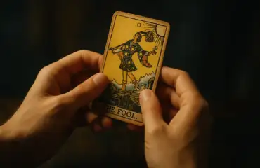 Руки начинающего таролога держат карту «Шут» из колоды Райдера-Уэйта в тёплом мягком свете
